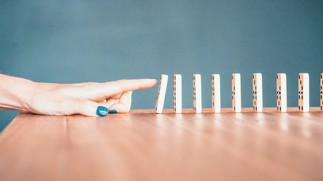 Un index s'apprête à pousser le premier domino d'une longue rangée alignée sur une table en bois.